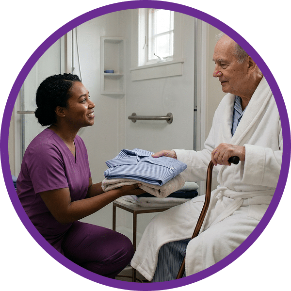 Nurse in purple scrubs hands folded clothes to an elderly man in a bathrobe, seated with a cane.